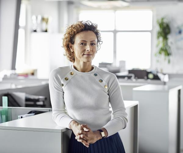 Businesswoman with hands clasped standing in an office and leaning against a table 