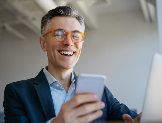 Older smiling businessman sitting in front of a computer and looking at his phone